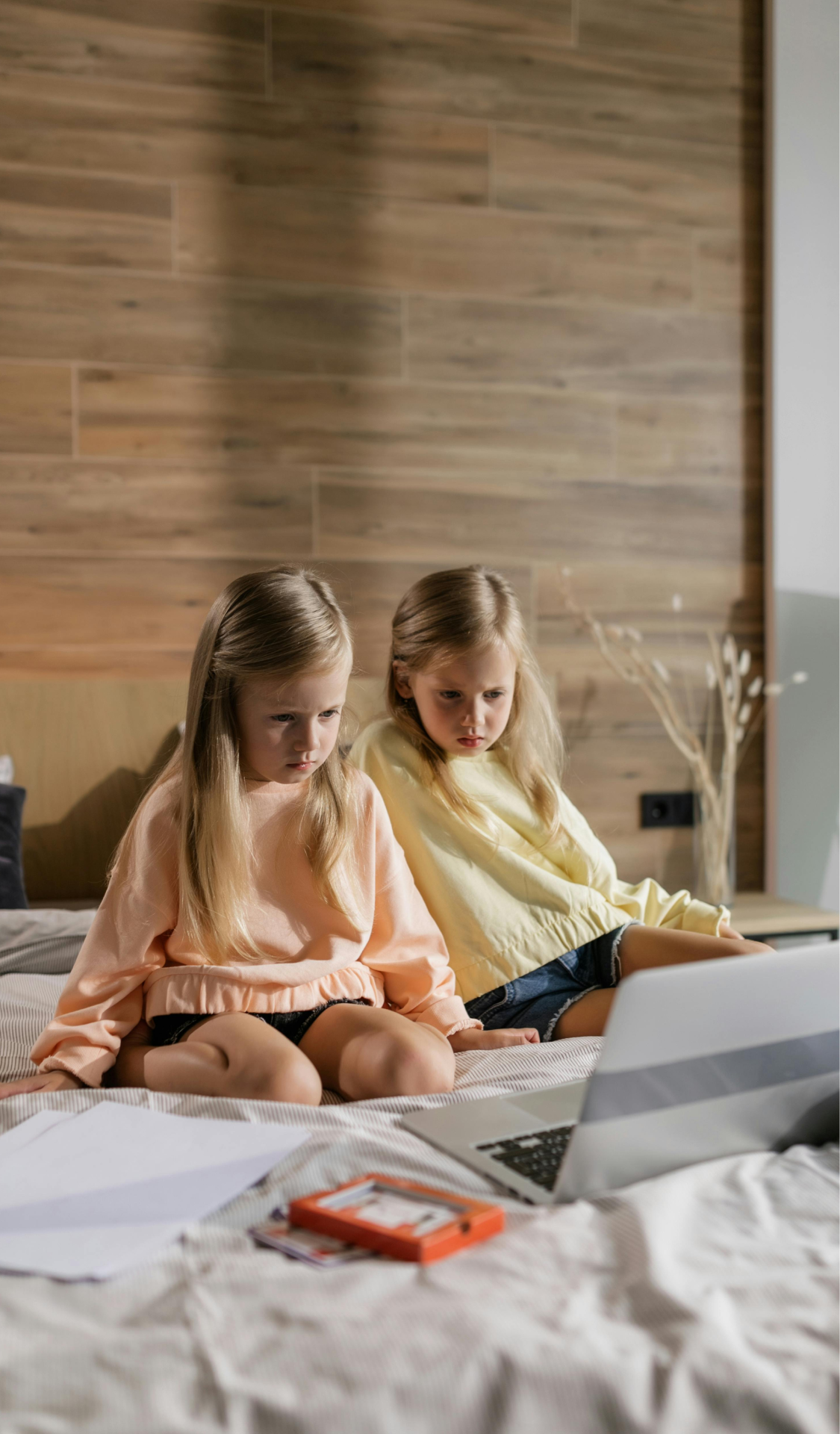 Two young girls in casual sweatshirts (peach and yellow) sitting on a bed with a laptop, papers, and an orange device, appearing to be engaged in online learning